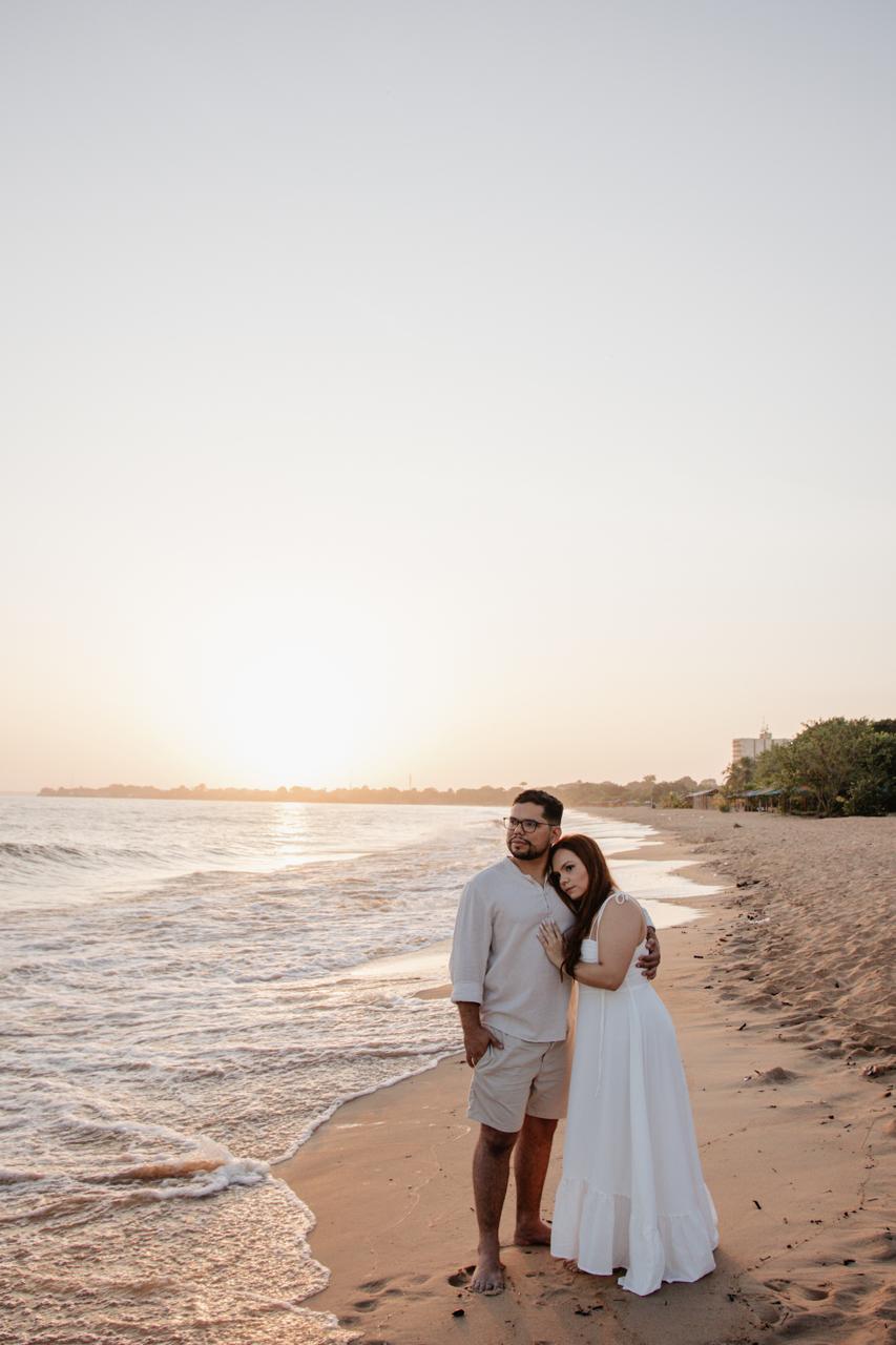 Aline e Artur abraçados na beira do mar ao pôr do sol